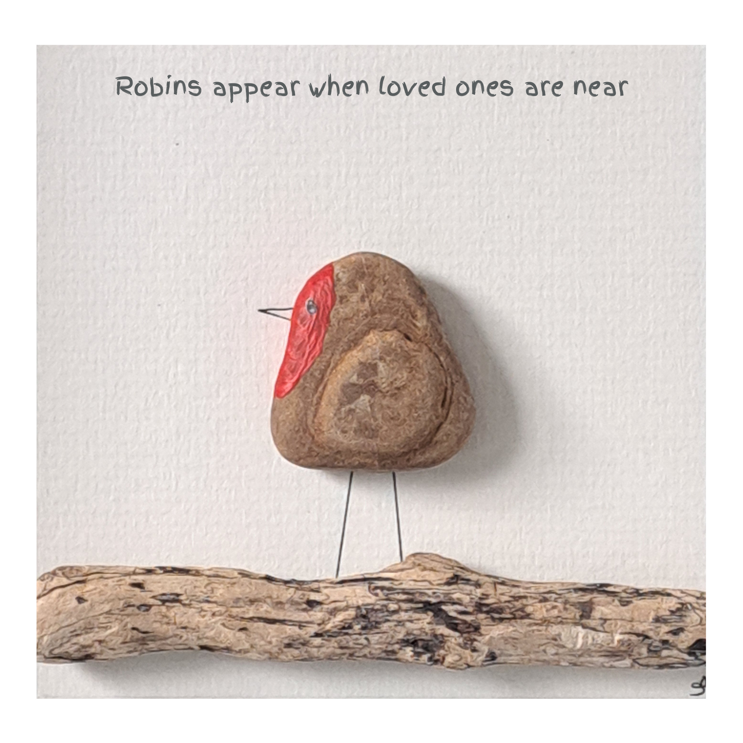 Decorative stone with natural wing markings and a red painted breast, placed on a white surface with text and a piece of driftwood.