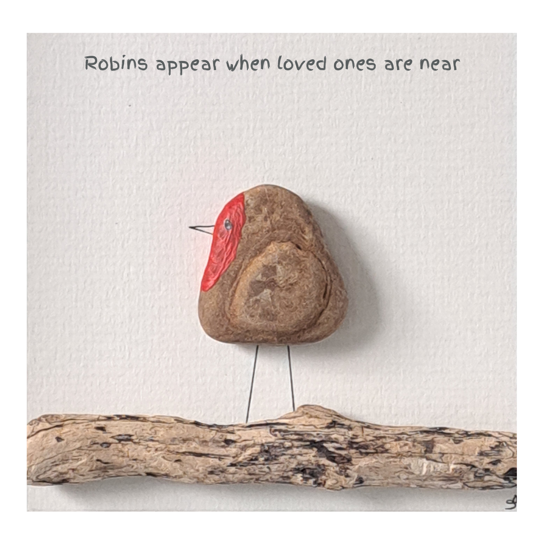 Decorative stone with natural wing markings and a red painted breast, placed on a white surface with text and a piece of driftwood.